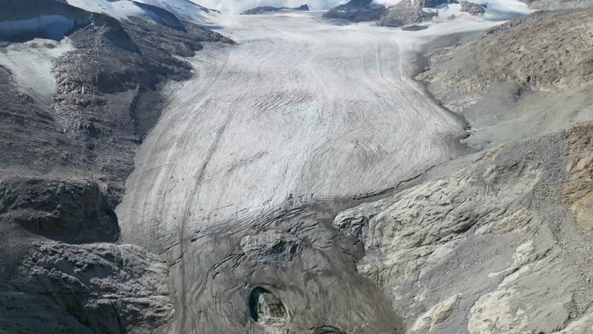 Aerial view of a glacier winding through rugged mountains, their peaks capped with snow under the sun, Brescia, BS, Italia.