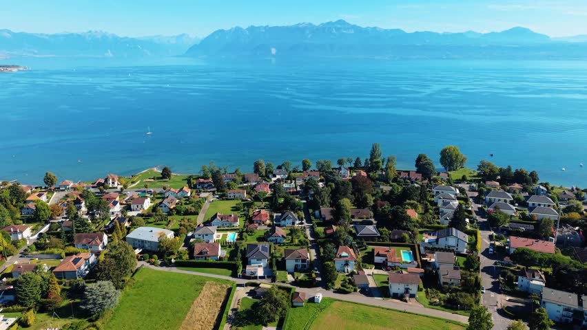 Aerial view of houses with pools nestled by the serene Lake Geneva, set against distant mountains and clear blue skies, Saint-Sulpice, Vaud, Switzerland.