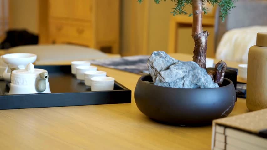 A slow pan across a wooden table reveals a traditional white ceramic tea set and a bonsai tree. The tranquil scene evokes a peaceful minimalist lifestyle.