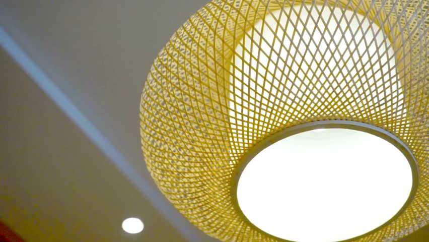 A slow pan of a modern, woven bamboo pendant lamp hanging from the ceiling. The stylish, eco friendly light fixture provides a warm and cozy ambiance.