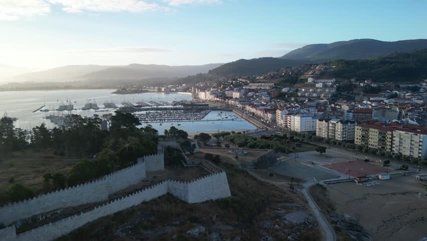 Aerial view of boats docked in the harbor with city buildings and mountains under a blue sky with white clouds, Baiona, Pontevedra, Spain.