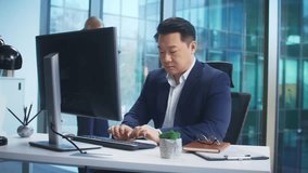 Positive Asian man in elegant suit smiling while working at computer in modern office. Typing report or managing data. Blonde female colleague standing in background using smartphone. - Powered by Shutterstock - Get 15% off with code: PIKWIZARD15