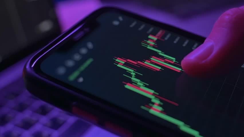 Close up of smartphone with trading app and candlestick charts in trader hand. Crypto investor analyzing cryptocurrency or equity market using mobile device. Neon light