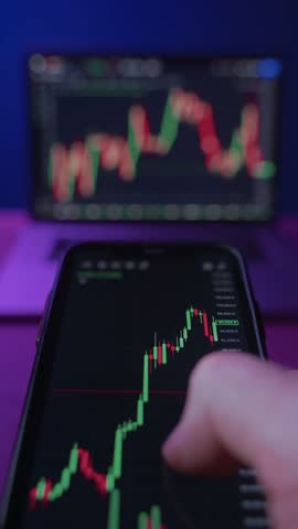Close up of trader holding smartphone, using trading application analyzing crypto and stock market trends at his neon lit office with multiple monitors on background. Vertical video