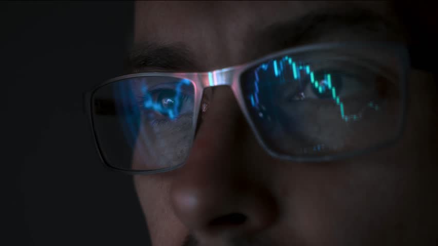 Close up view of focused trader wears eyeglasses looking at pc screen with stock market charts reflection, working online late night. Man analysing crypto market. Selective focus on eye
