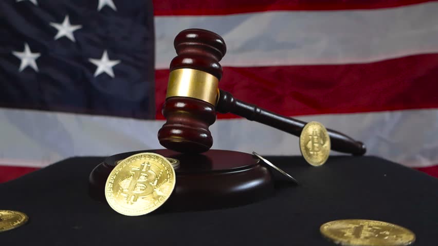 Brown wooden court hammer with bronze decorations placed next to golden shiny Bitcoin cryptocurrency money coins that are all in front of a United States of America flag while video orbits around them