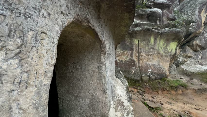 A large, carved archway leads into a dark cave, with the ancient stone walls covered in historical carvings and a mossy rock formation visible in the background