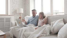 Father and daughter sit on sofa with laptop, smiling and waving to someone during online video call in bright cozy home. Concept of family bonding, communication, parenthood, connection and love - Powered by Shutterstock - Get 15% off with code: PIKWIZARD15