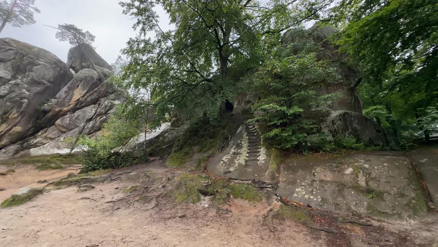 A view of large, mossy rock formations with lush green trees, featuring a set of stairs carved directly into the stone, all under a cloudy sky