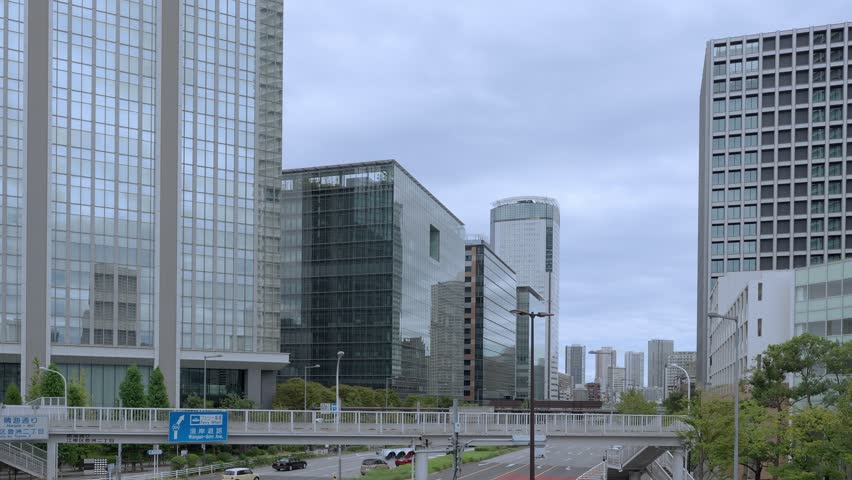 A low-angle shot of a Tokyo cityscape with a variety of modern glass skyscrapers and buildings against an overcast sky.