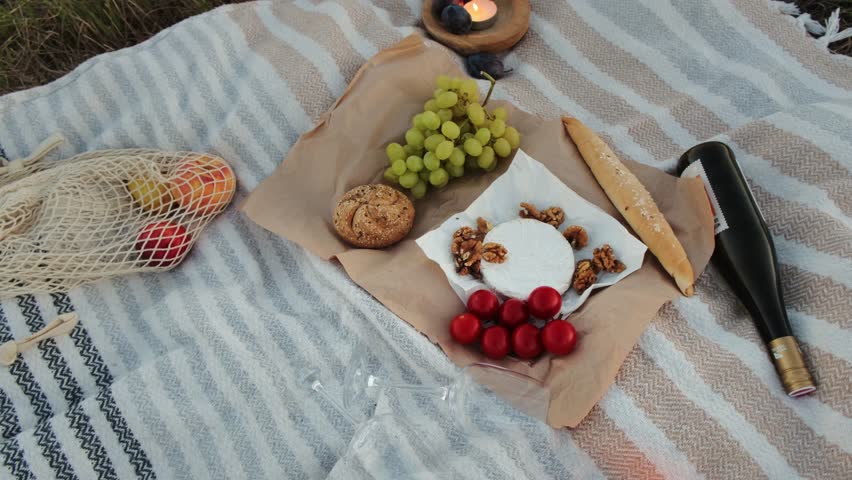 Close-up of a cozy picnic on a light blanket in Vienna’s vineyards. Camembert with walnuts, tomatoes, grapes, bread, wine, fruits, and candle create warm, romantic vibes.