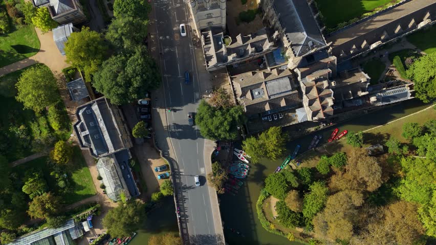 Aerial top-down drone shot of Magdalen College, Oxford. The flight moves diagonally over the Gothic tower, main building, and green courtyard, with dramatic long shadows.