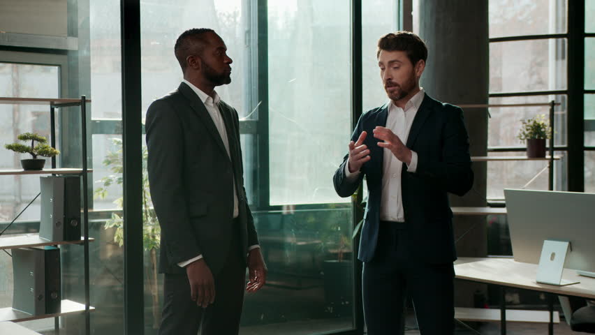 Two serious multiracial business men team businessmen talking in office company meeting multiethnic colleagues partners discuss work job deal Caucasian African American employers talk negotiation