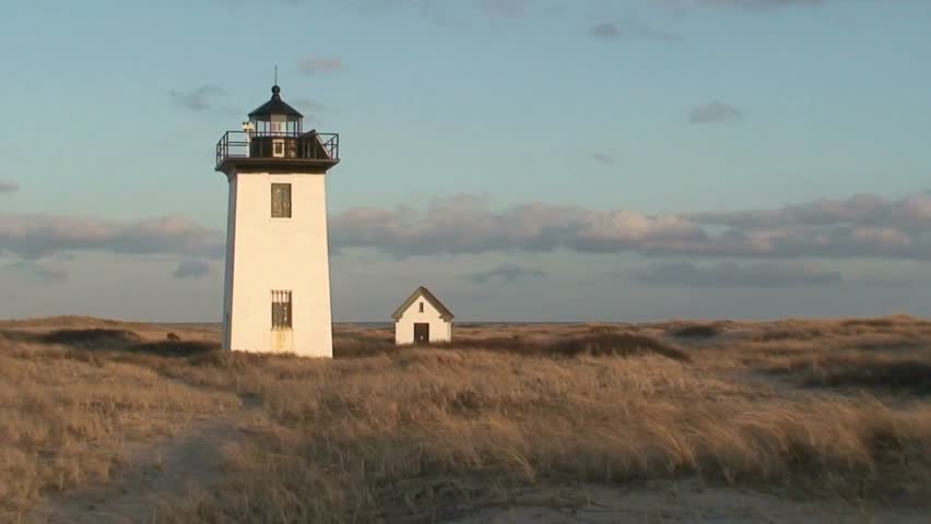 A white lighthouse stands tall in a dune landscape at sunset