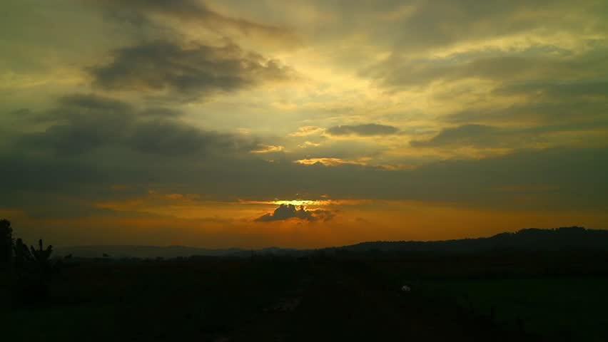 A dusk view with a sky filled with dark clouds and the light of the setting sun shining behind it, creating the silhouette of mountains or hills in the distance.