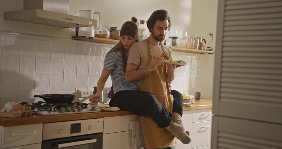 Happy girl hugging her boyfriend and sitting on kitchen countertop after cooking breakfast together and having breakfast in cozy kitchen at home In the morning