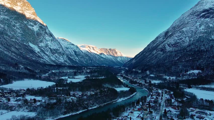 aerial view captures the serene winter landscape of Oppdal, Norway. The footage showcases the charming town nestled within a picturesque valley, flanked by majestic snow-covered mountains under a clea