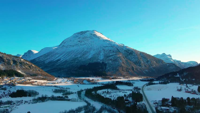 An aerial view captures the serene winter beauty of Oppdal, Norway, showcasing snow covered mountains, a picturesque valley with a winding river or road, and scattered buildings of the town under a cl
