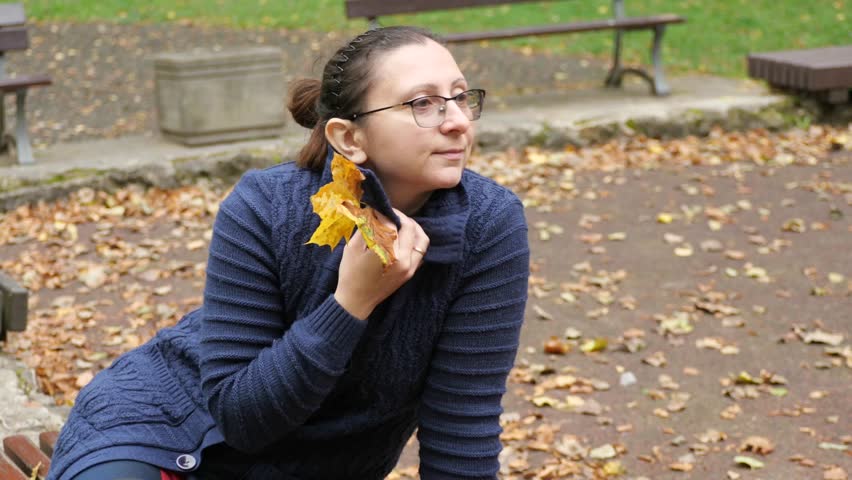 A woman sitting in an autumn park - A woman in a cozy blue sweater sits on a park bench, holding a yellow autumn leaf. The ground is covered in fallen leaves, creating a perfect, peaceful scene