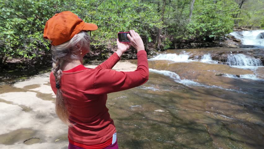 A mature woman enjoys hiking among stunning waterfalls and lush greenery in Babcock State Park, West Virginia. Capturing her adventure with a smartphone.