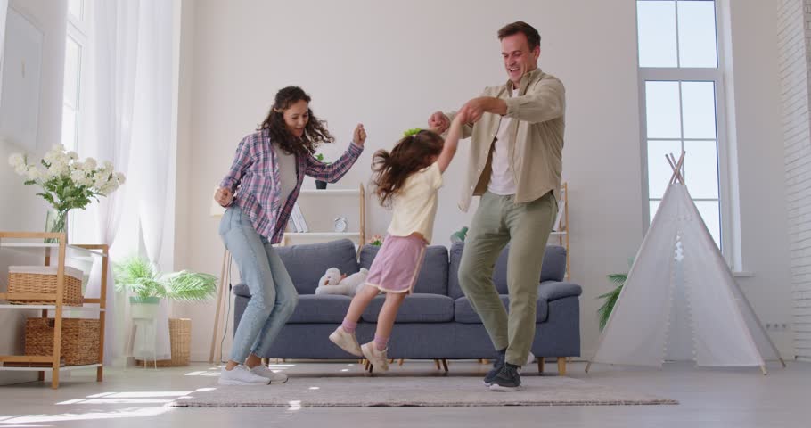 Family dancing with daughter in livingroom at home. Parents spin the child by the arms, stepping near the sofa as daylight fills the room. Celebrates joyful family bonding at home together.