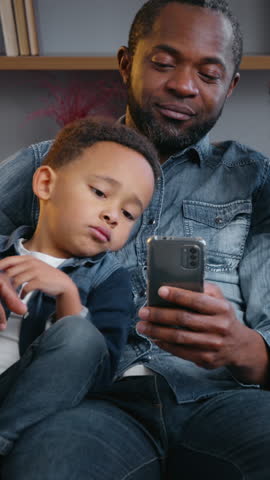 Happy African American family using mobile phone app at home together father with sons looking in smartphone gadget addict smiling talking browsing social media kids children boys with dad on couch