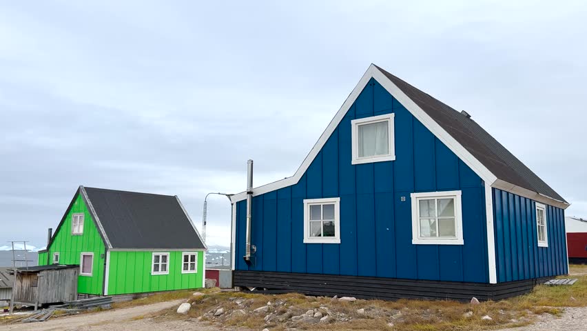 Colorful houses sit on rocky terrain near calm water in Greenland. The backdrop features impressive snow-capped mountains and floating ice. The clear blue sky enhances the tranquil setting.
