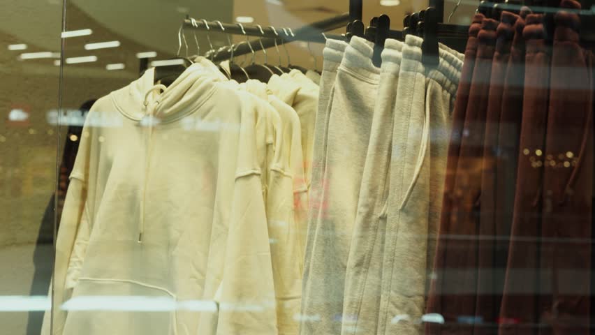hoodies and sweatpants in neutral tones hanging on racks inside a clothing store, viewed through a glass window