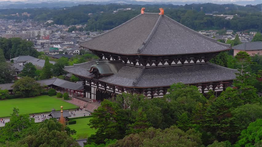 Aerial view of Nara, Japan, showcasing Todai ji temple's intricate roof, lush green hills, and a mix of historical and modern structures with smooth motion.
