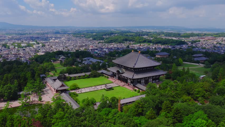 Aerial view of Todai ji Temple in Nara, Japan, surrounded by greenery and pathways. The town and mountains appear under a partly cloudy sky.