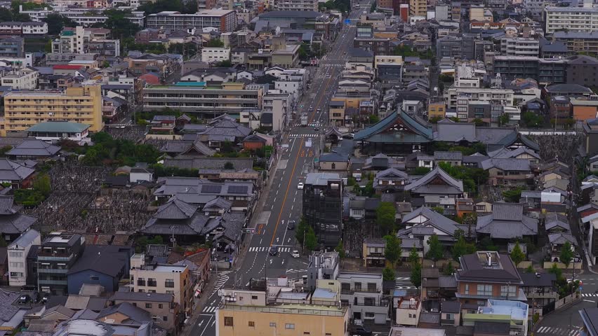 Aerial view of Kyoto, Japan, showing a central road, traditional rooftops, a temple, and modern buildings, with green hills in the background.