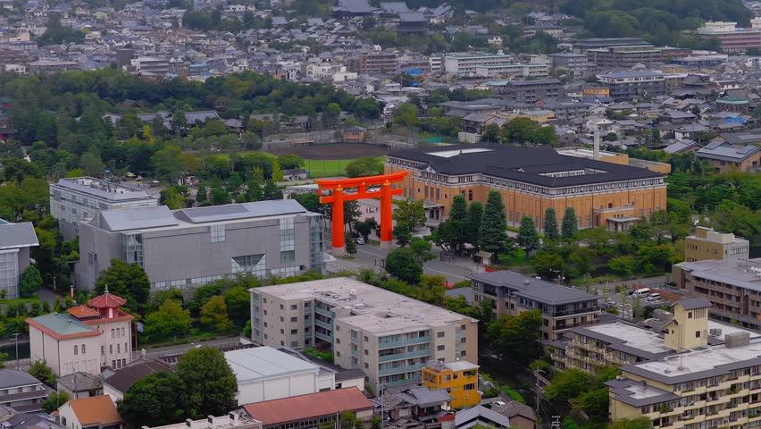Aerial view of Kyoto, Japan, featuring a large orange torii gate, traditional rooftops, a temple in the hills, and a mix of urban and natural scenery.