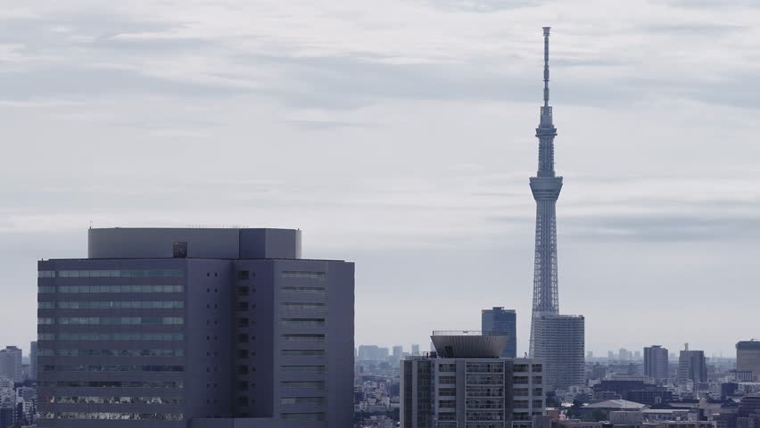Aerial panning view of Tokyo, Japan, featuring the Tokyo Skytree, modern high rises, and smaller structures under a cloudy sky.