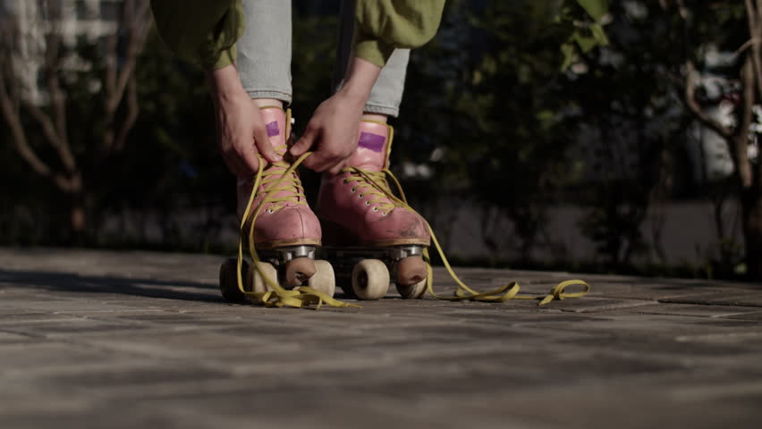 Woman hands tie yellow laces of pink roller skates on paved road