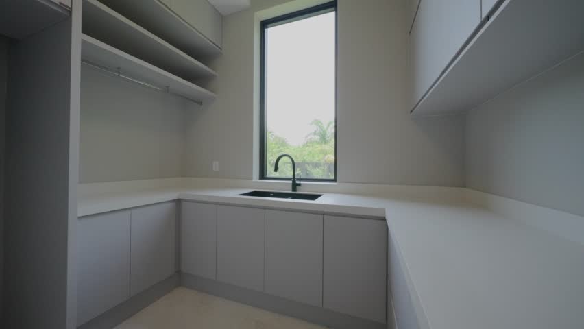 Modern kitchen or utility room with light gray handle less cabinets and white L shaped countertop. Black faucet and sink sit below tall window that brings in natural light