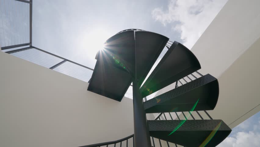 Black spiral staircase viewed from below leading up toward the sky. Staircase is surrounded by white walls and metal railings. Sun shines through steps