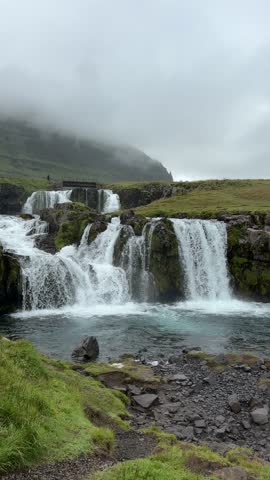 Powerful waterfall cascades over mossy rocks into clear blue pool with misty mountains and cloudy sky in Grundarfjörður, Iceland. Filmed July 31 2025.