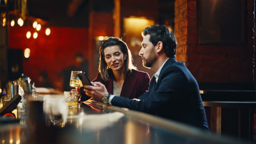 Man woman drinking alcohol sitting at modern pub counter using smartphone. Flirting people clinking glasses saying toasts at evening date atmosphere. Charming couple chatting in quiet stylish lounge