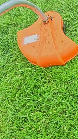 Bright orange trimmer head actively cuts green grass in slow motion, blades moving through fresh lawn as particles scatter creating dynamic garden work scene