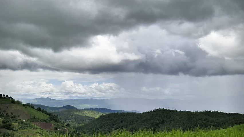 A stunning aerial view captures vibrant green terraced fields on hillsides. Dark storm clouds gather above, with heavy rain visibly falling in the distance over the majestic mountains. 