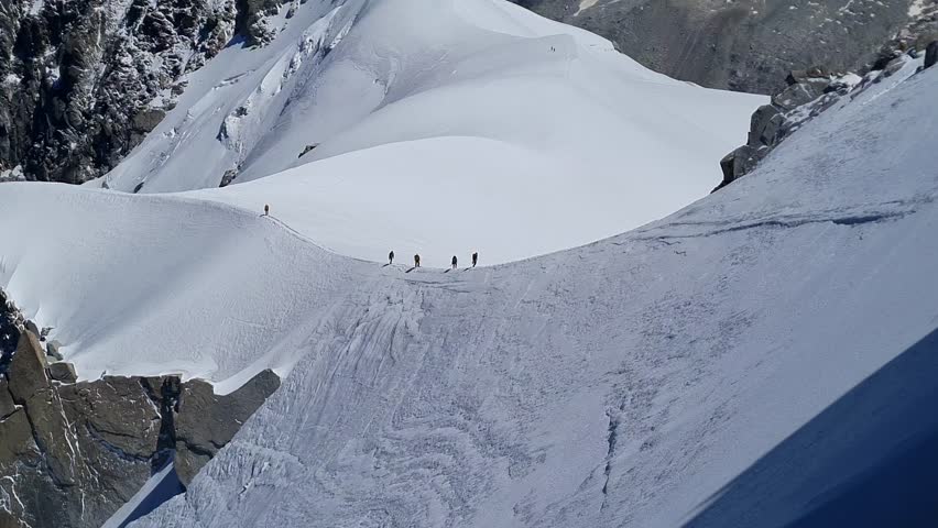 A group of five mountaineers slowly trekking along a narrow, snow-covered mountain ridge.