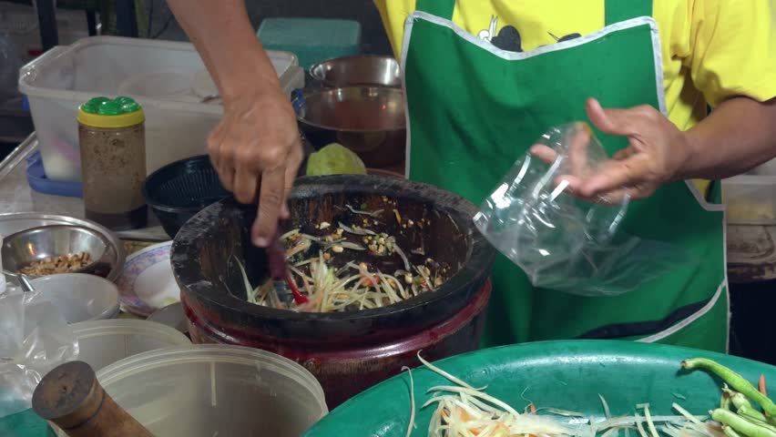 A street vendor skillfully prepares authentic som tam papaya salad, pounding fresh shredded papaya with chili, lime, fish sauce, and vegetables in a mortar for a flavorful Thai street food dish.