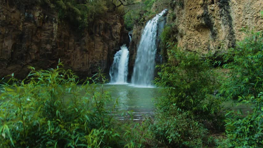 Green natural pool with triple waterfalls cascading from high cliffs, tilt up drone shot at Turbinas Santa Cruz, Jalisco, Mexico