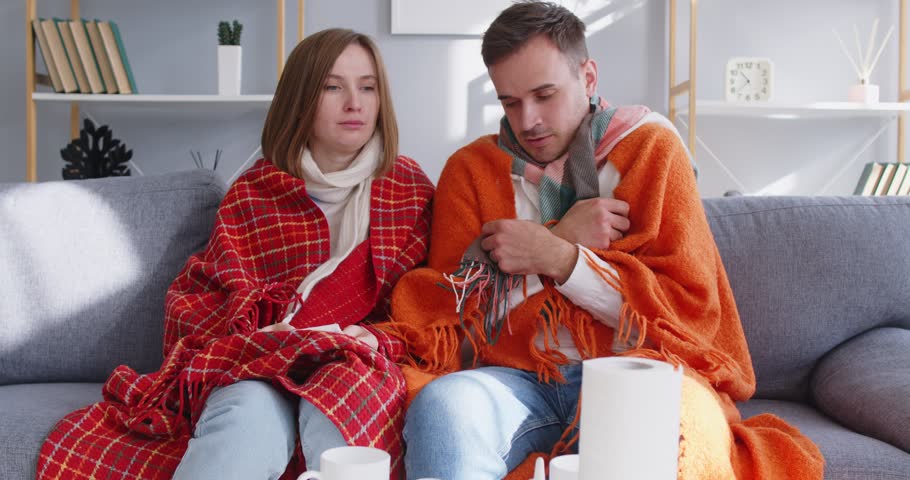 Young couple covered in blankets sitting on sofa at home with seasonal flu or cold. Sick unhappy man with woman sneezing suffering from runny nose and feeling unhealthy with influenza. 4k video.