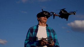 Flying fpv drone pilot controls quadcopter in blue sky wearing goggles and holding controller for outdoor hobby flight, drone pilot monitors flight path while quadcopter hovers near him in afternoon - Powered by Shutterstock - Get 15% off with code: PIKWIZARD15