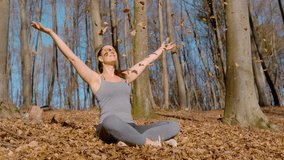PORTRAIT, SLOW MOTION: Content, young woman sits peacefully on a sunny day in the forest, with her eyes closed and arms outstretched as she enjoys the sun and the autumn leaves falling around her. - Powered by Shutterstock - Get 15% off with code: PIKWIZARD15