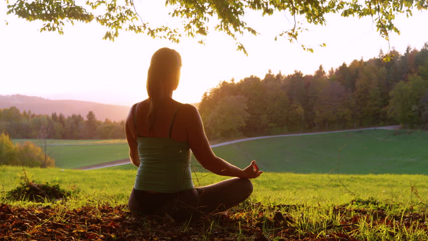 LENS FLARE, SILHOUETTE: Woman sits in a peaceful, meditative position on a grassy hill, facing a beautiful golden sunset. Setting sun shines from behind her, emphasizing serene, peaceful atmosphere.