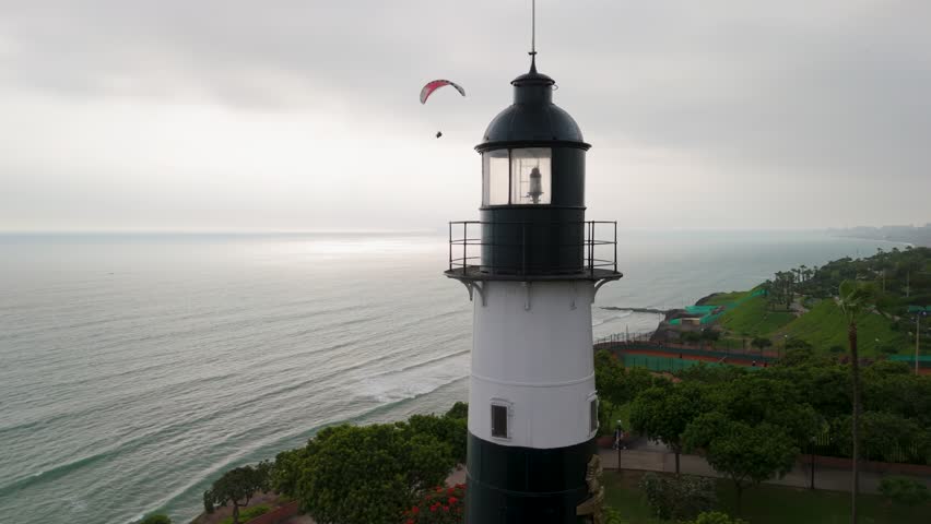 A stunning aerial parallax shot circles the historic Miraflores Lighthouse (Faro La Marina) in Lima, capturing a paraglider soaring over the Pacific Ocean with the beautiful coastline in view