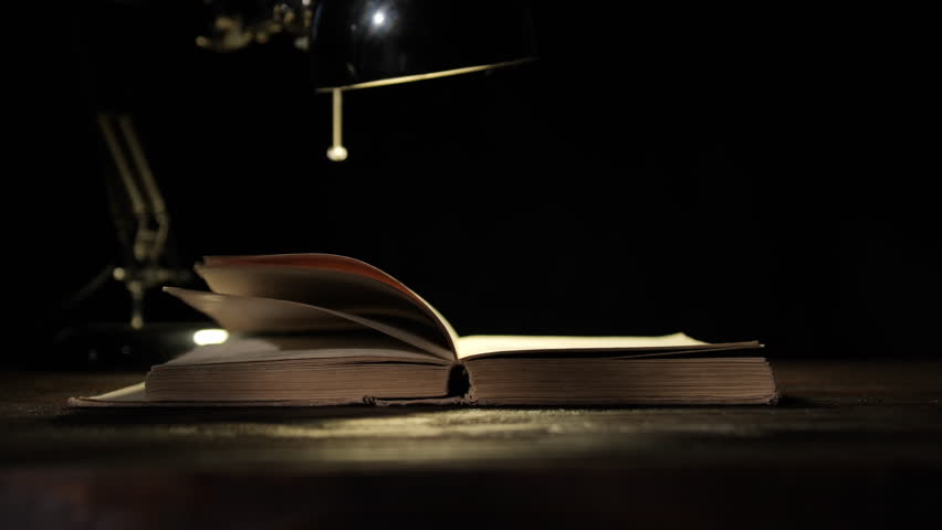 Pages of old book turning under light of table lamp on black background close-up