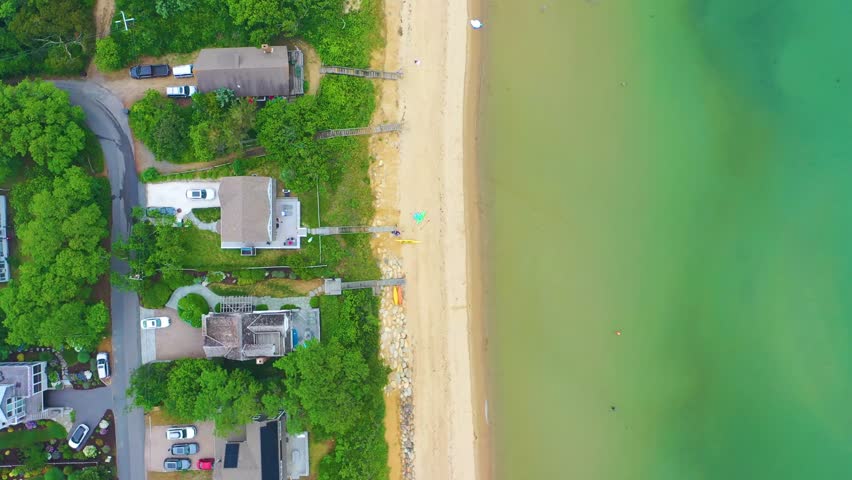 Overhead drone video of Cape Cod neighborhood with beachfront houses, sandy shoreline, calm waters, and green landscape, with residential roads bordering the coast.